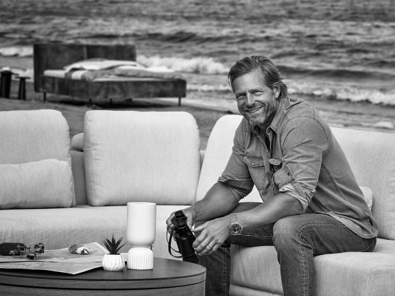 Ein Mann sitzt lächelnd auf einem Sofa am Strand, mit einem Bett im Hintergrund und Wellen im Meer. Er genießt die ruhige Atmosphäre.