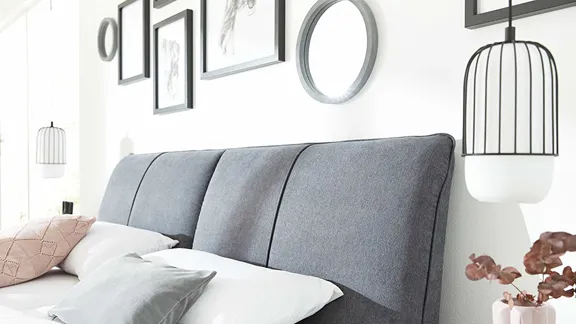 Detailansicht der grauen, gepolsterten Bettkopfstütze mit Kissen vor heller Wand mit Bildern und Lampen.